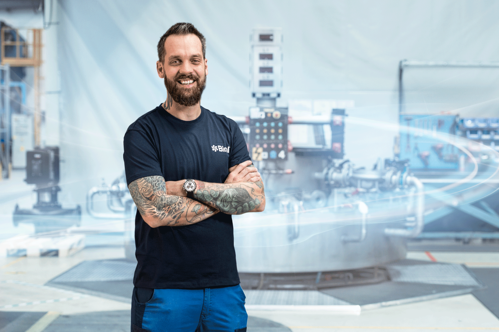 Mann mit tätowierten Armen in der Produktionshalle vor Maschine.