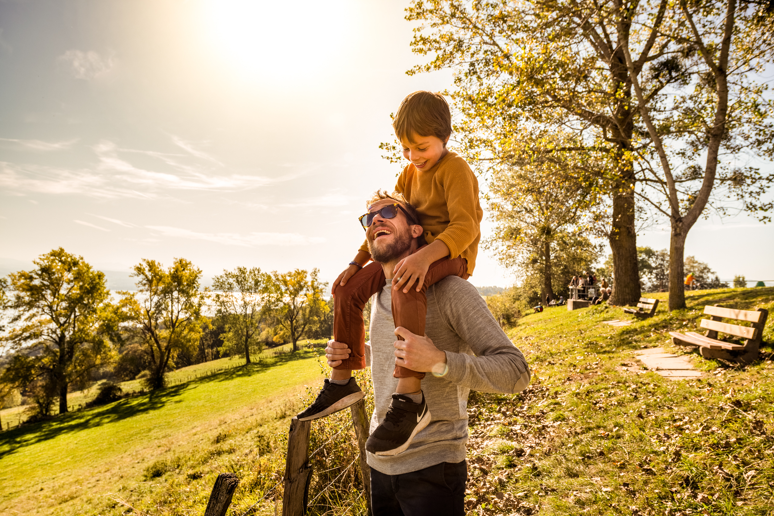 Vater mit Sohn im Park in Murten. Mann mit Pulli und Sonnenbrille trägt Kind auf den Schultern in der Natur. Vater und Kind spielen zusammen im Freien auf einer Grünen Wiese mit Sitzbank und Bäumen.