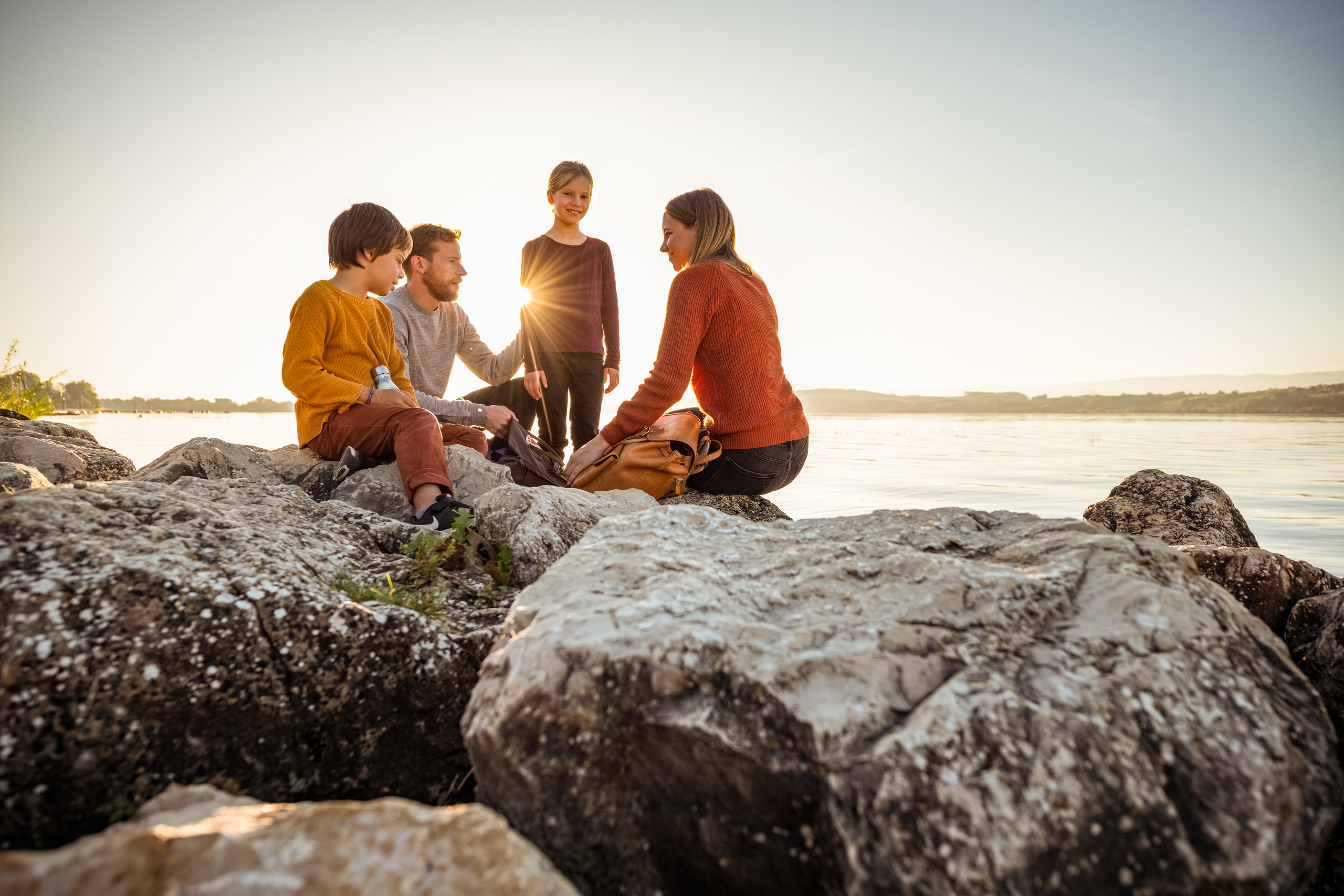 Familie am Murtensee bei Sonnenuntergang. Mutter mit Vater und Kind sitzen auf Felsen am See.