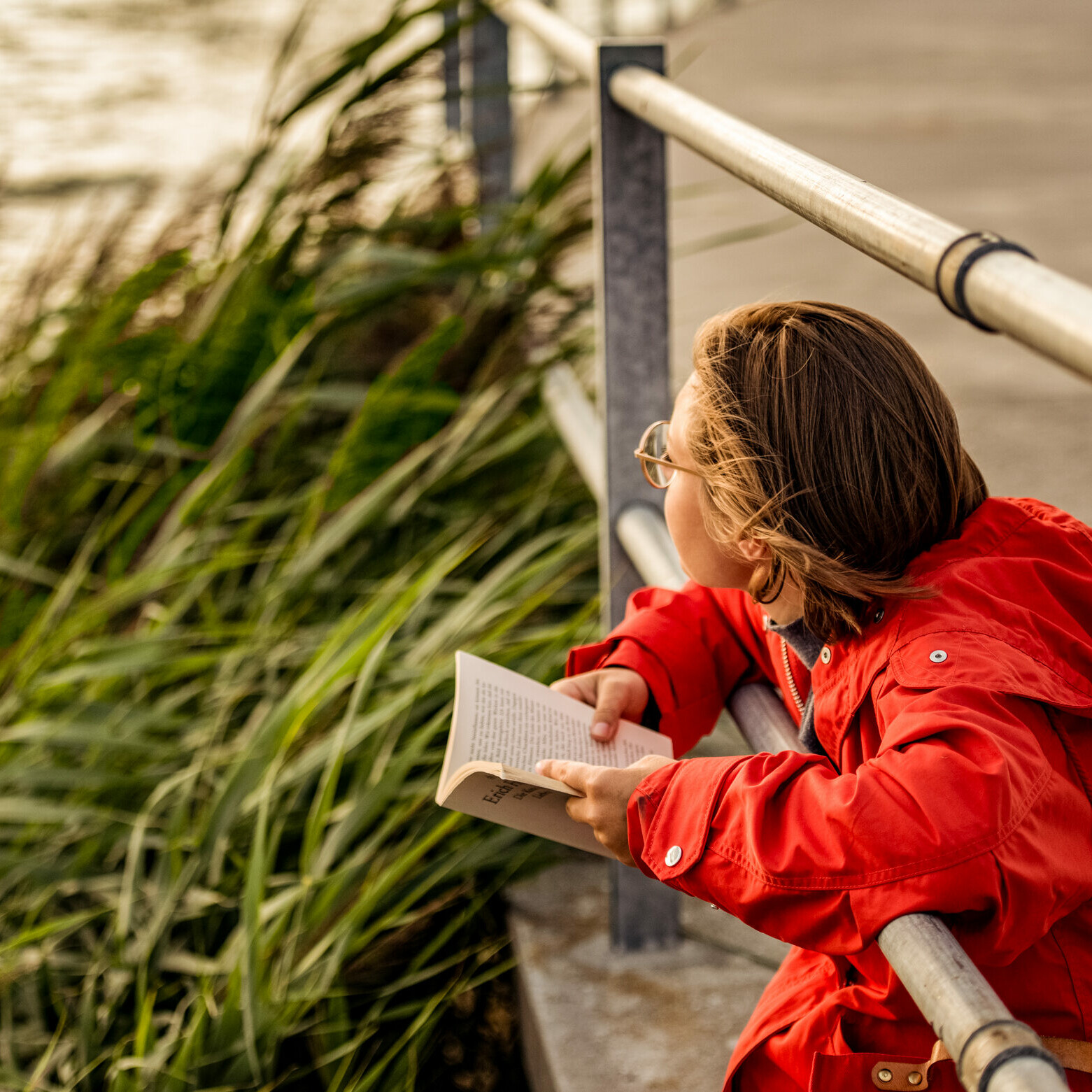 Frau sitzt mit Buch am Rand vom See in Murten. Junge Frau mit rotem Regenmantel sitzt am Seeufer und liest ein Buch.