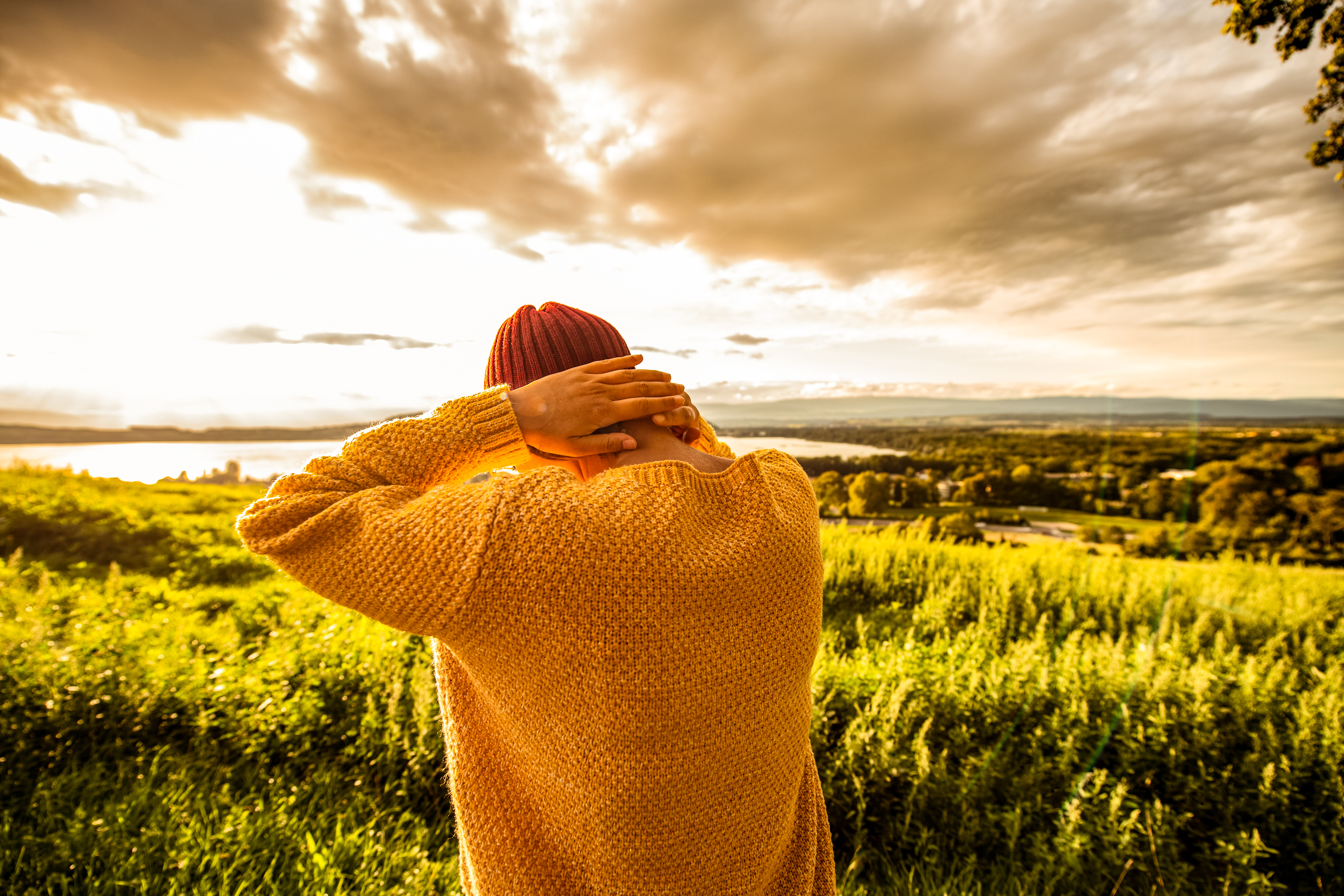 Junge Frau mit roter Kappe und gelben Pullover auf einem Hügel. Junge Frau mit gelbem Pullover steht auf einer grünen Wiese, starke Wolken ziehen auf.