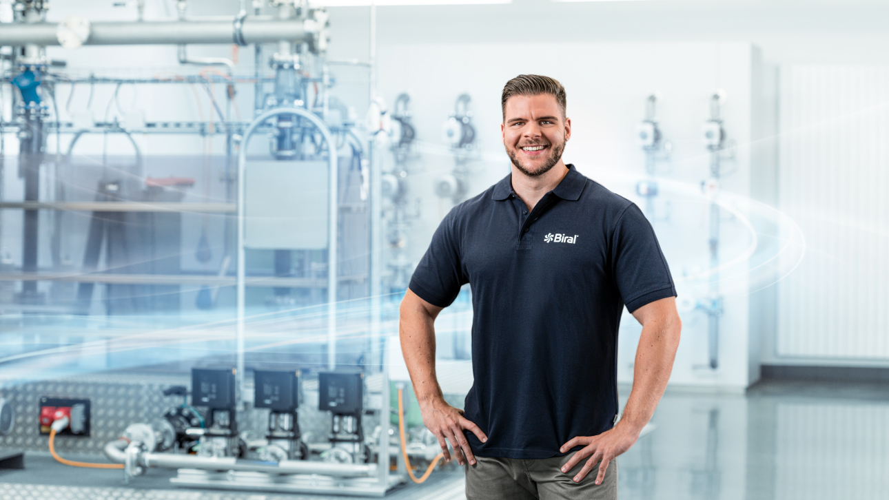 Mann in Fabrik, steht vor grosser Maschine. Imagebild mit blauen Akzenten für Biral AG. Junger Mann mit dunkelblauem T-Shirt lächelt bei der Arbeit.