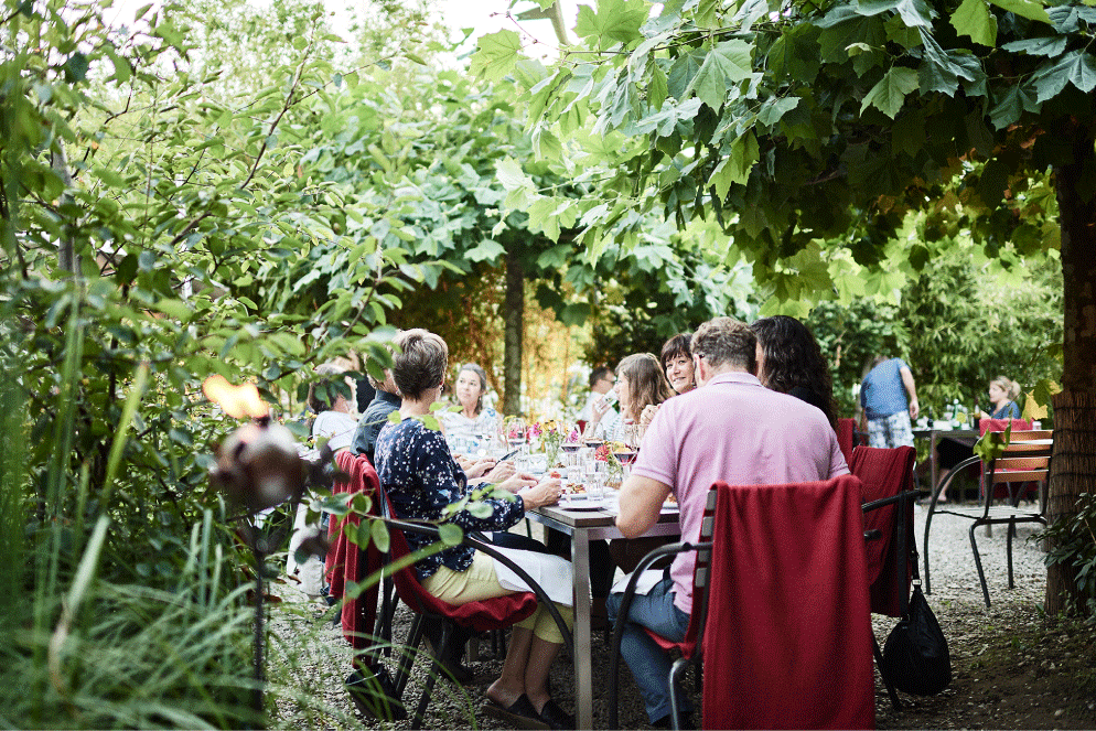 Menschen sitzen auf der Gartenterasse. Gartenterasse mit vielenBäumen und grossem Tisch.