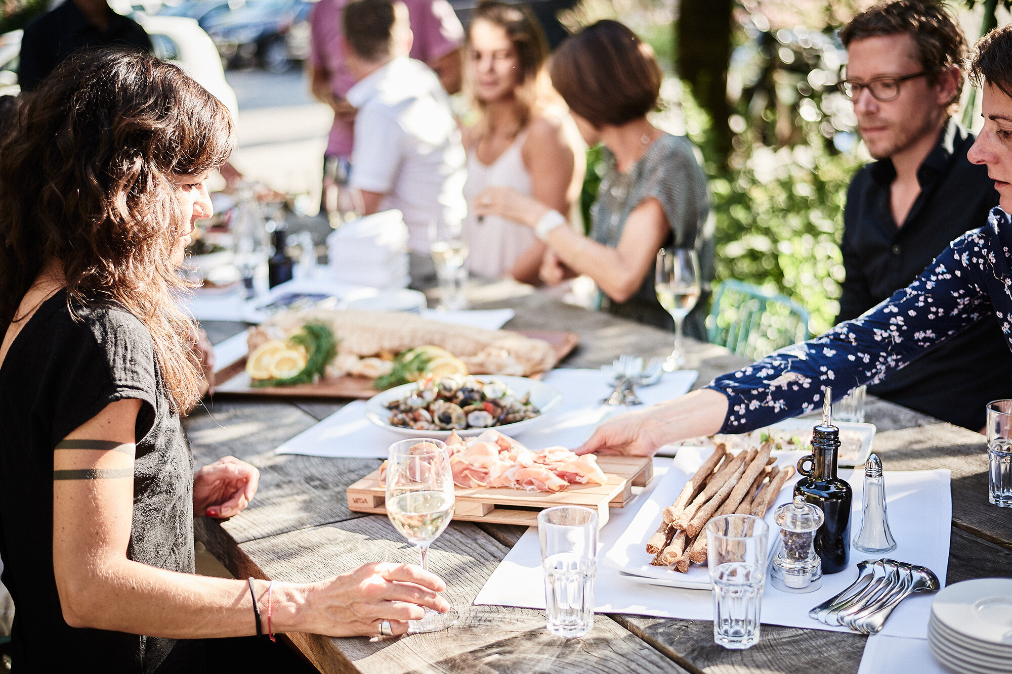 Menschen sitzen im Garten um grossen Tisch und essen gemeinsam. Frau streckt sich nach Fleischplatte.