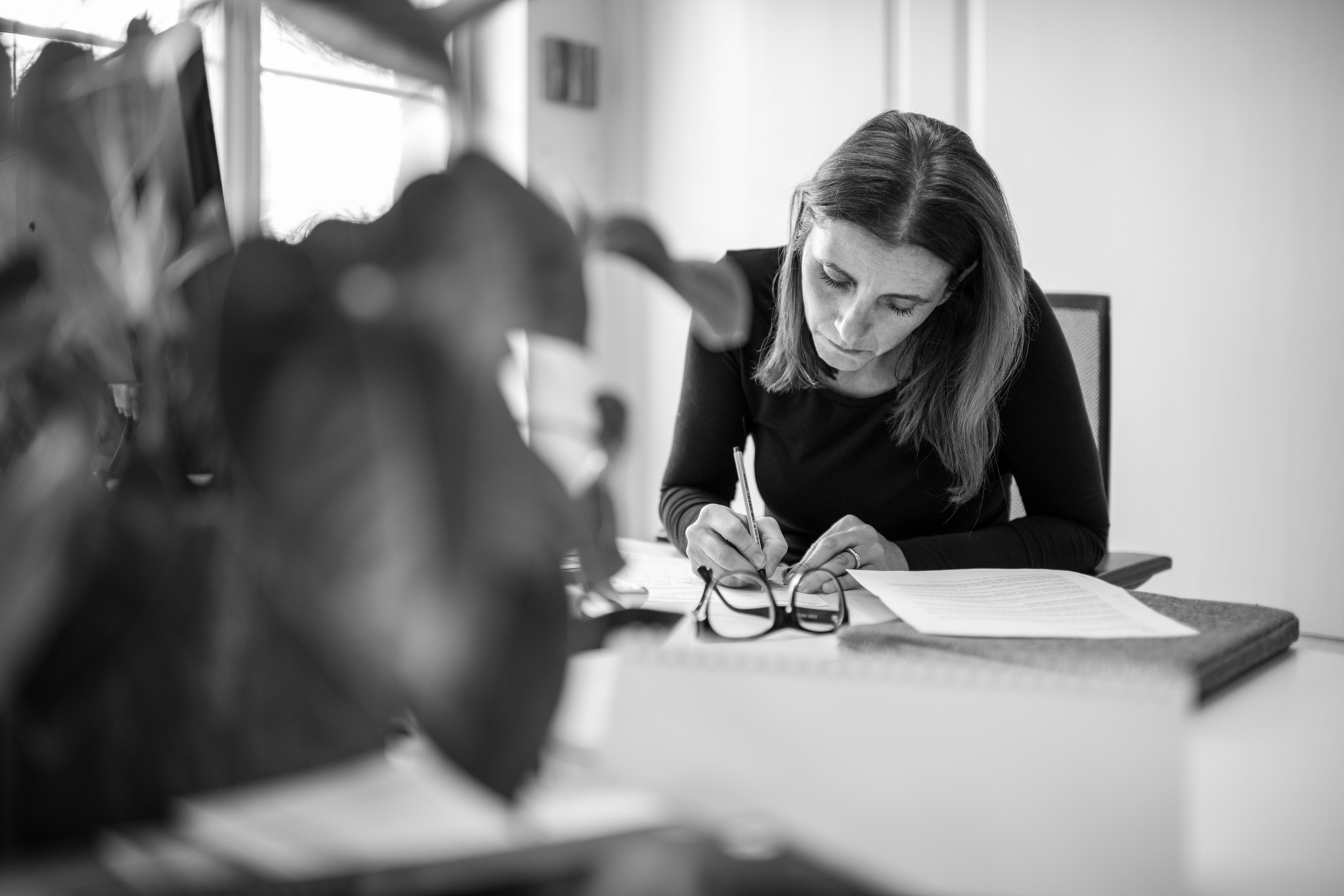 Cristina Laillard sitzend am Tisch bei der Buchhaltung. Buchhalterin notiert sich etwas. Brille liegt auf dem Tisch im Vordergrund steht eine Pflanze.