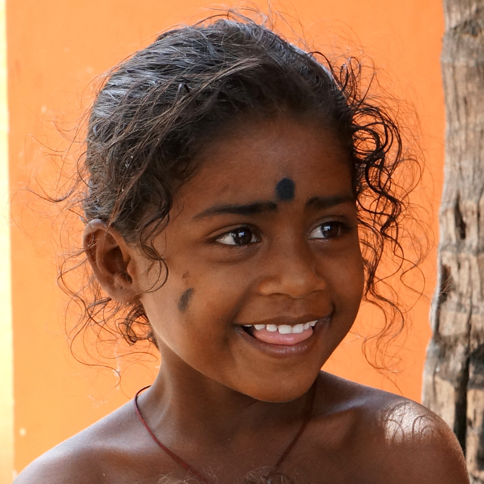 Indisches Mädchen lächelt vor Orangefarbenem Haus in Indien. Kleines Mädchen mit Mal auf der Stirn am Grinsen in Indien.