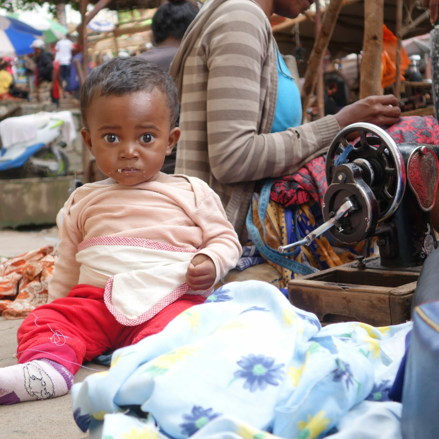 Kleiner Junge sitzt in einem Slum am Boden und isst etwas. Frauen im Slum am arbeiten mit kleinem Buben mit roter Hose und Lachsfarbenen Pullover