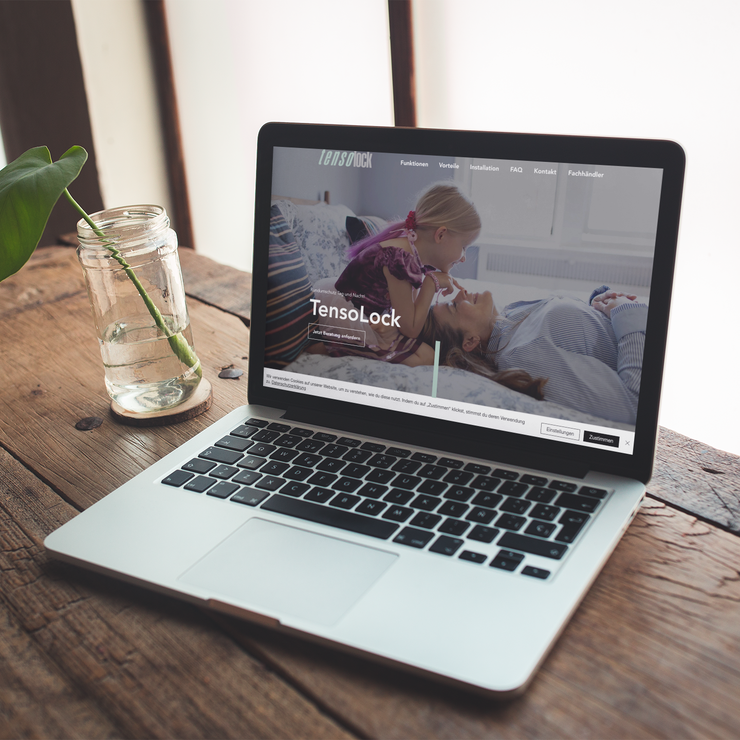 Mac Book steht auf Holztisch zu Hause. Daneben steht ein Glas mit einem Blatt
